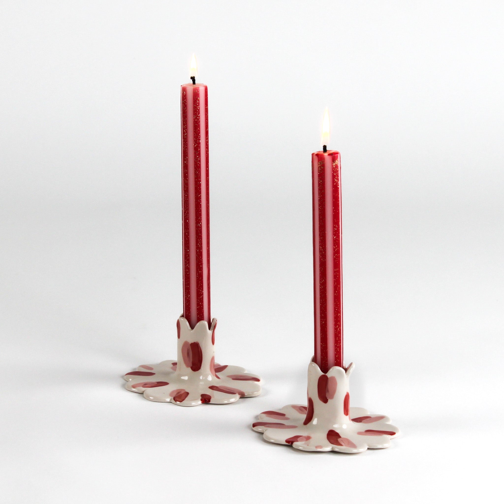 Two lit red taper candles displayed in white ceramic scalloped candle holders with hand-painted red brushstroke accents on a light background.