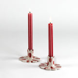 Two lit red taper candles displayed in white ceramic scalloped candle holders with hand-painted red brushstroke accents on a light background.