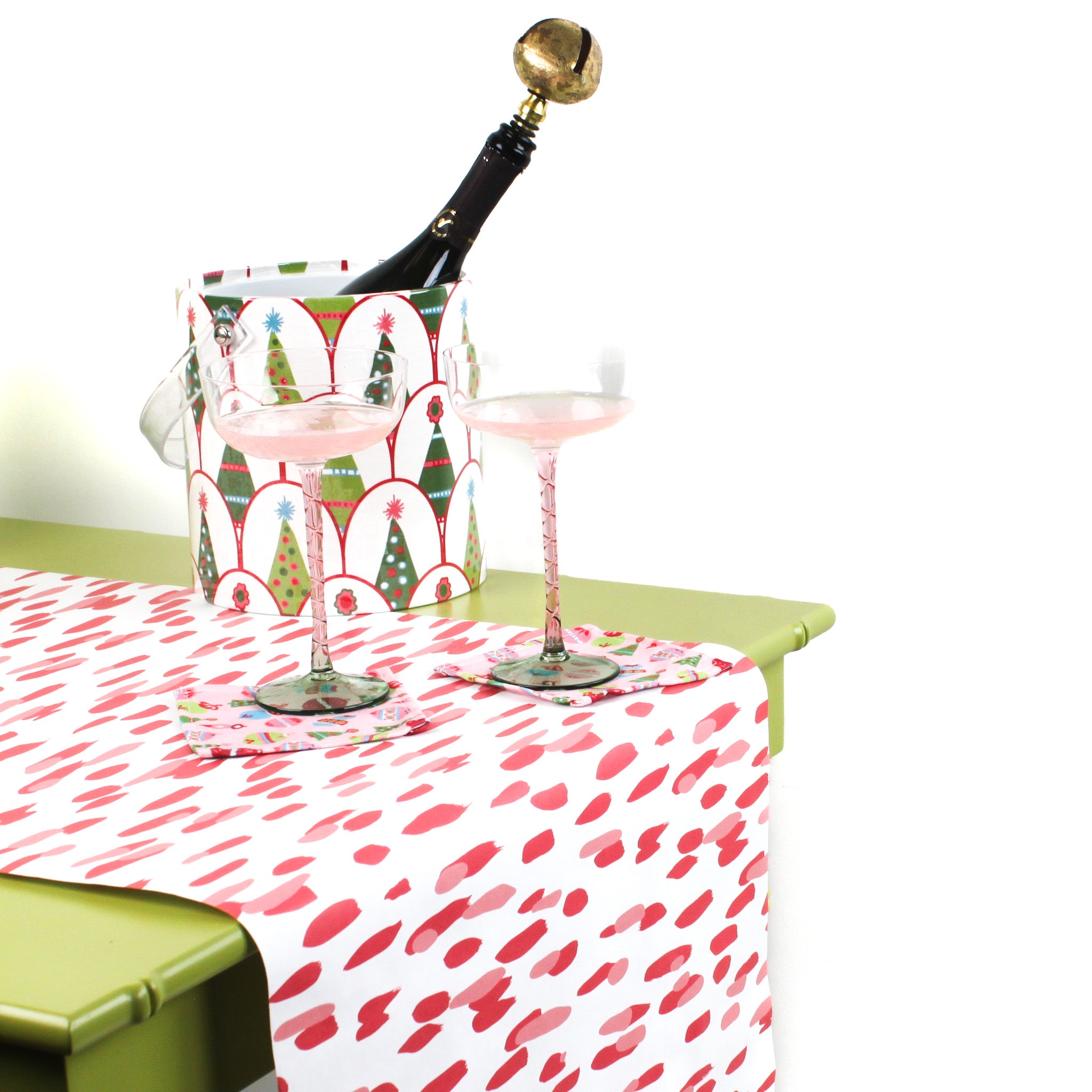 a green table covered with A pink and white paper table runner with a brushtroke pattern on a white background with 2 pink glasses and an ice bucket with wine on it.