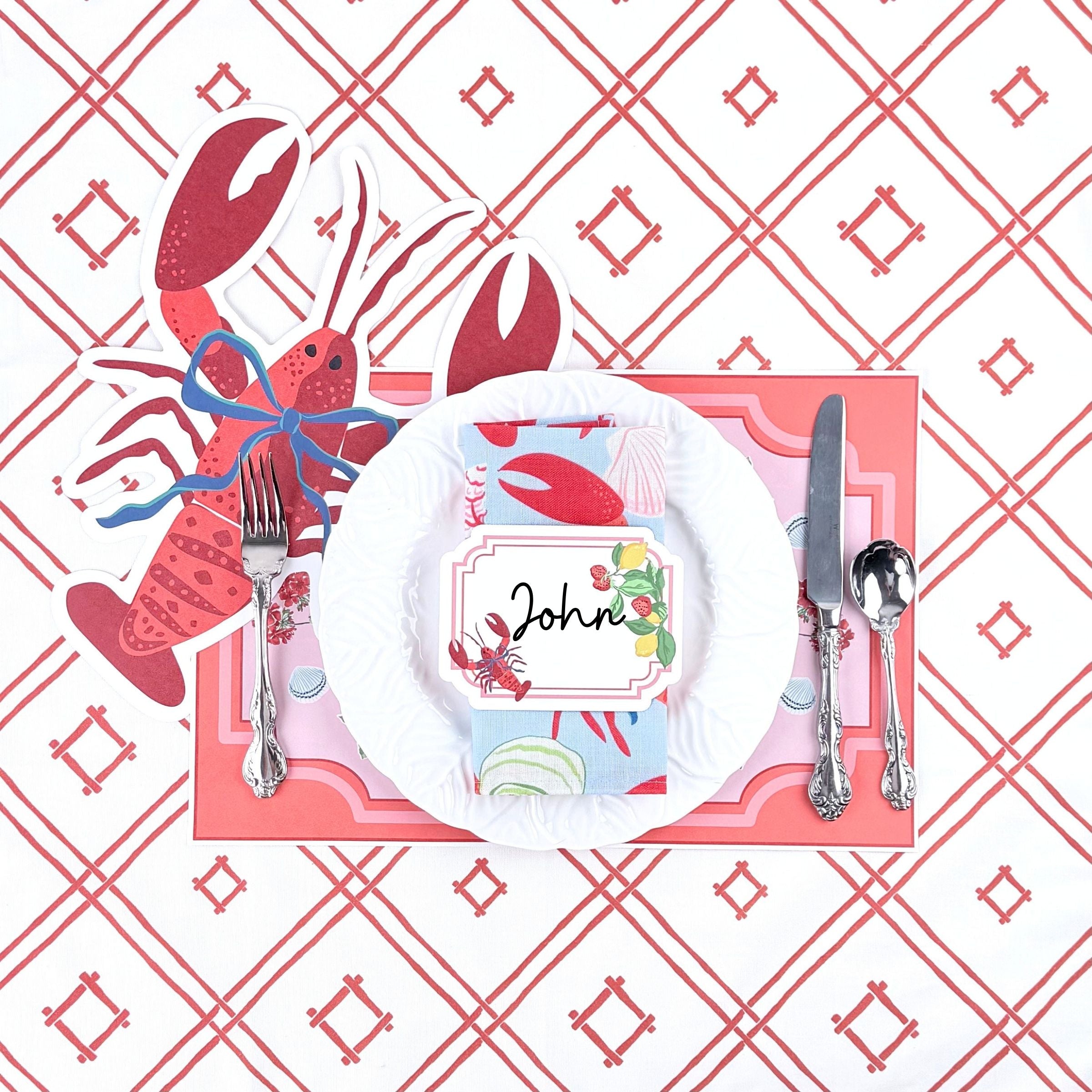 Coastal themed table setting featuring a lobster place card, lobster shaped placemat, white dinner plate, and coral trellis patterned background.
