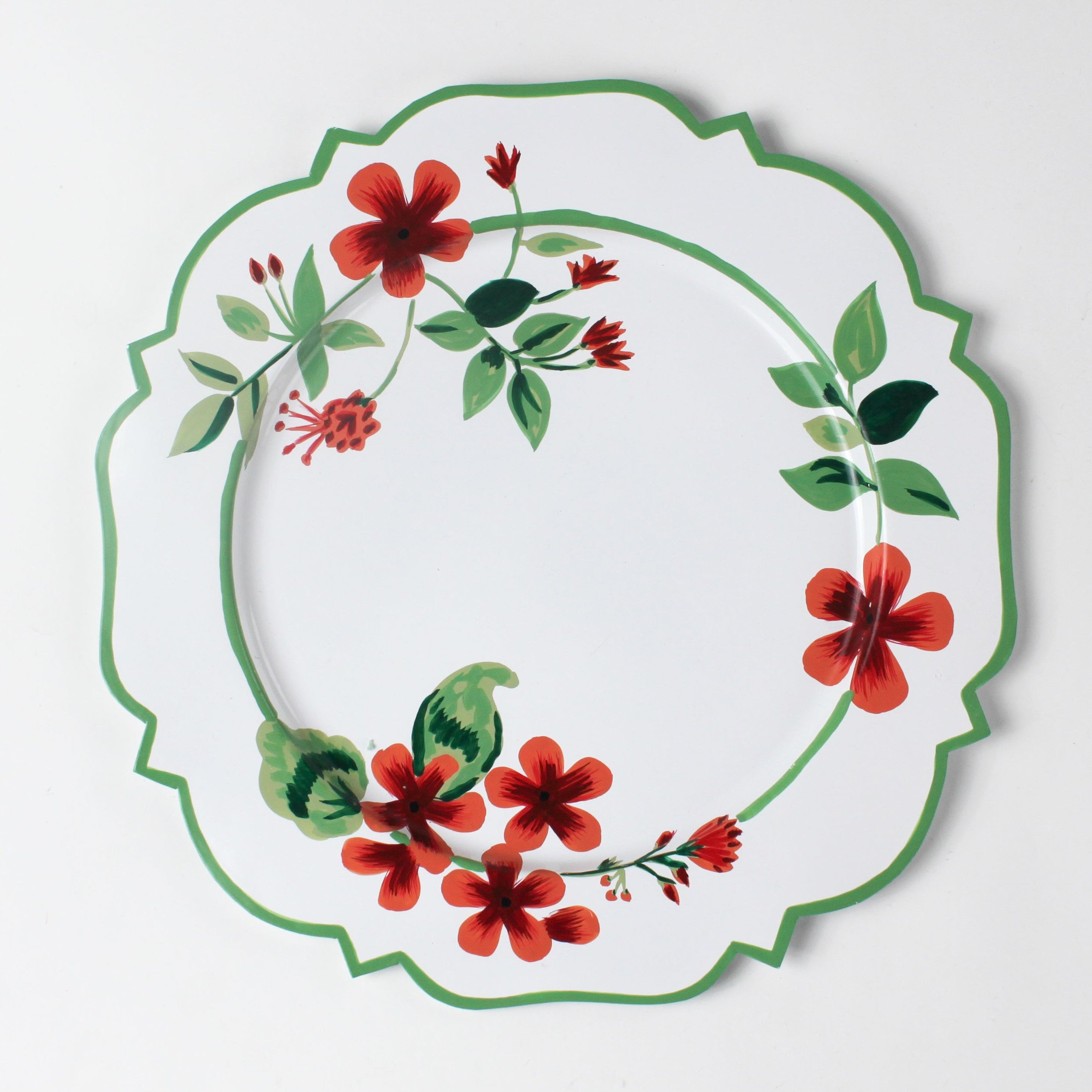 A white tole metal floral charger plate decorated with red geraniums with a green border against a white background. 