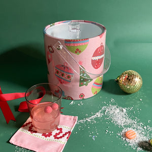 A pink ice bucket decorated with ornament motifs surrounded by cocktails and christmas decorations. 