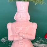 a close up shot of a nutcracker shaped pink ceramic christmas vase against a green, christmassy background. 