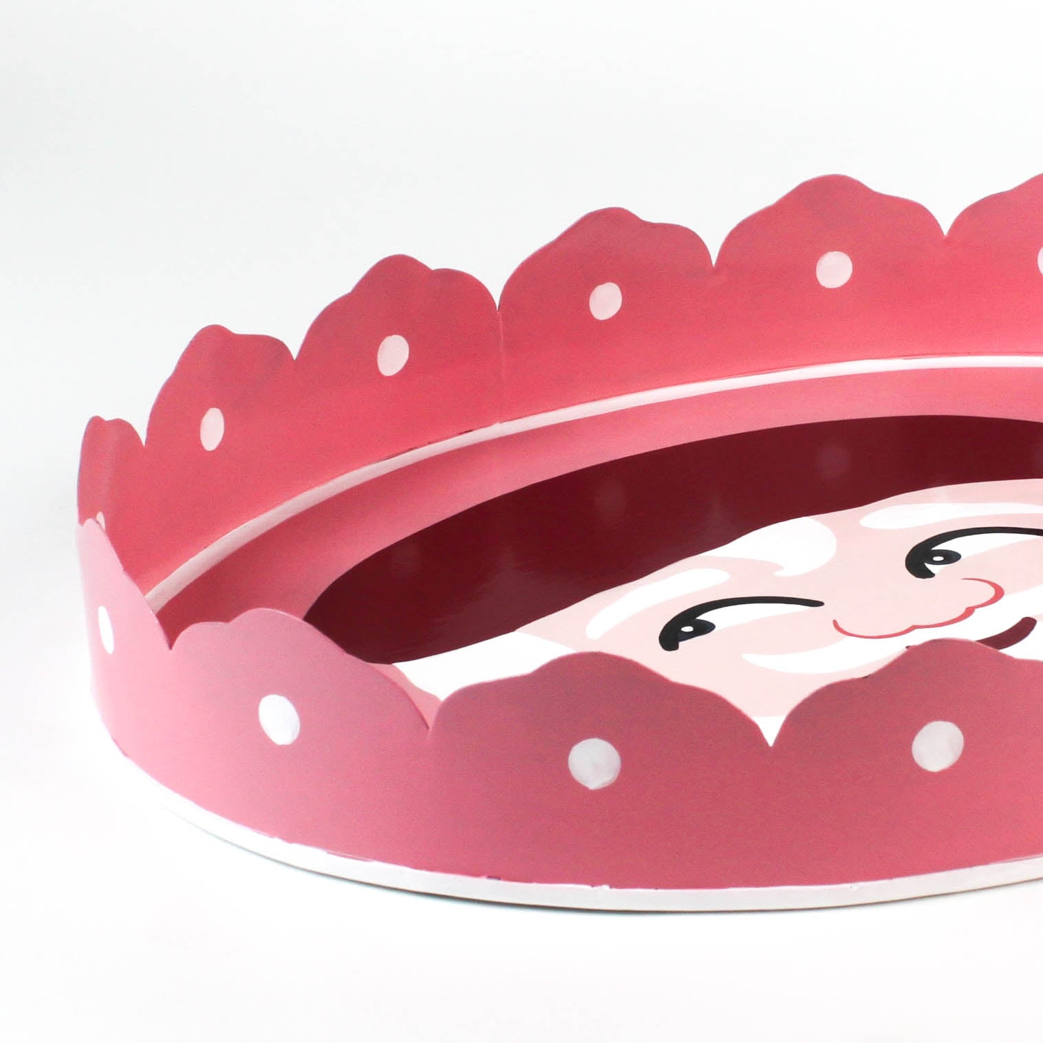 close up look of a Hand-Painted Metal Christmas Santa Tray featuring a cheerful Santa Claus face with a red hat on a pink circular background with scalloped edges.