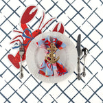 A styled coastal table setting featuring a lobster shaped paper placemat, white plate, nautical napkin, and silver utensils on a blue trellis patterned table surface.