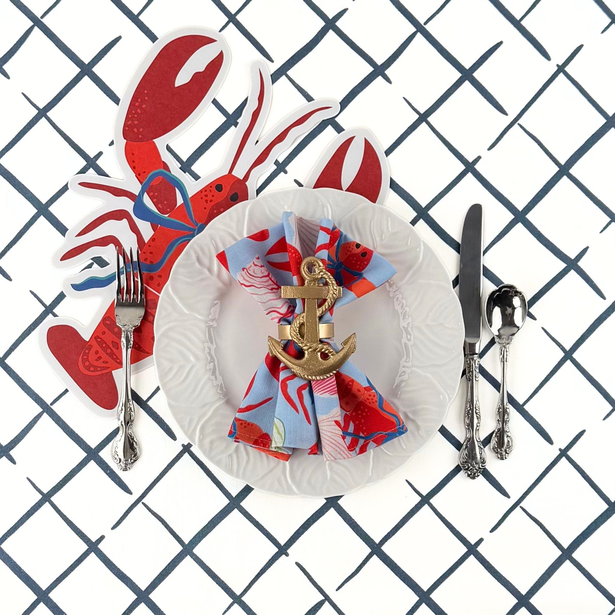 A styled coastal table setting featuring a lobster shaped paper placemat, white plate, nautical napkin, and silver utensils on a blue trellis patterned table surface.