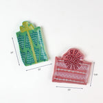 Two small ceramic plates shaped like gift boxes, one green with teal tree patterns and one pink with red ribbon designs, shown with dimensions around 3.5"–5.25" wide.