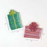 Two small ceramic plates shaped like gift boxes, one green with teal tree patterns and one pink with red ribbon designs, shown with dimensions around 3.5"–5.25" wide.