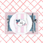 A styled table setting featuring a blue trellis paper placemat with a red ditsy floral border, white plate, pink napkin, and decorative place card.
