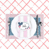 A styled table setting featuring a blue trellis paper placemat with a red ditsy floral border, white plate, pink napkin, and decorative place card.
