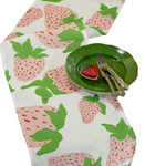 a strawberry fabric table runner designed by madcap cottage with motifs of large pink and green strawberries with green stem and a green plate resting on top.