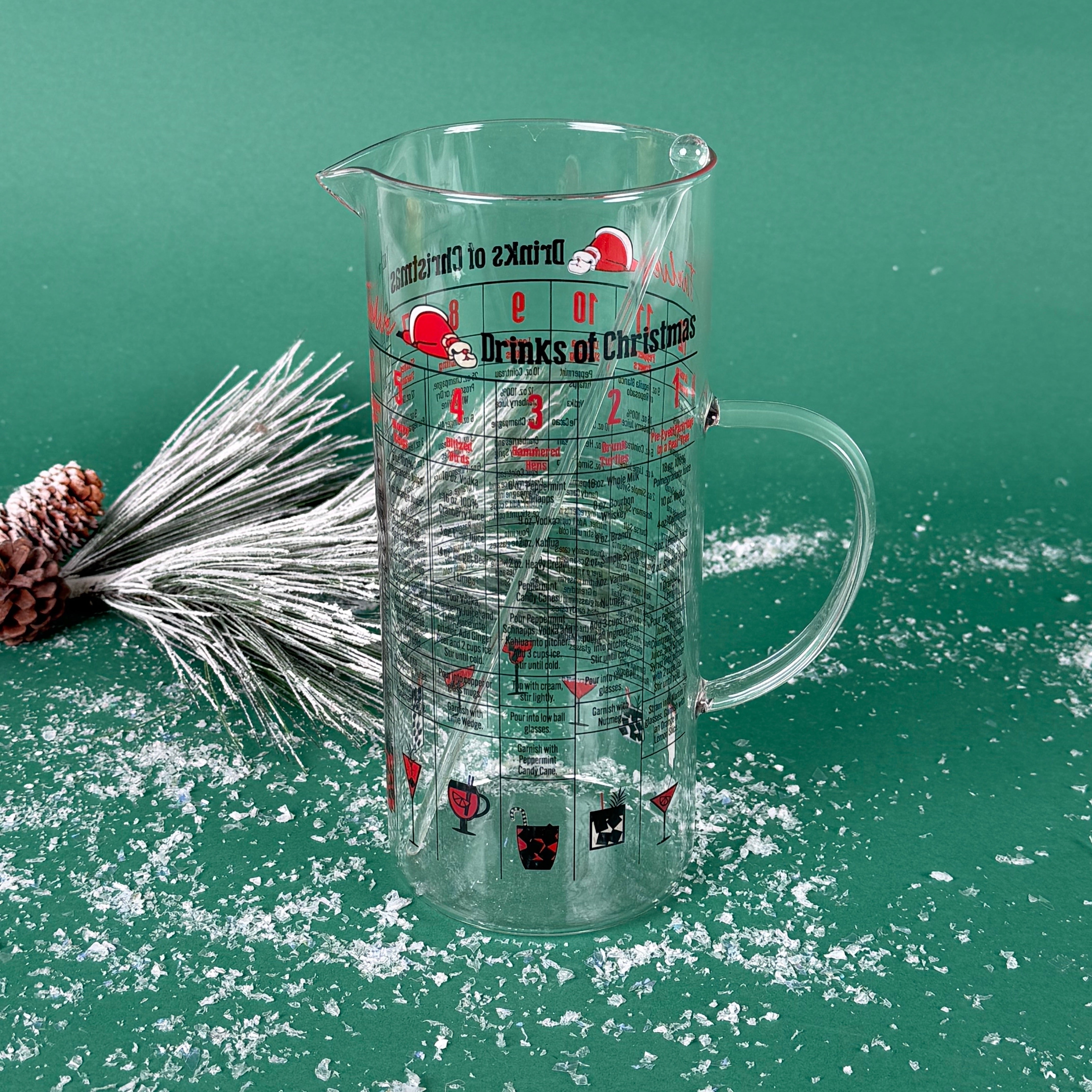 Clear holiday recipe pitcher labeled “Drinks of Christmas,” filled with a light pink drink, displayed on a snowy green background with frosted pine décor