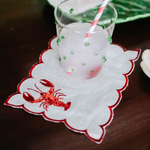 a white lobster napkin with an embroidered orange lobster motif and red scalloped edges with a glass resting on top