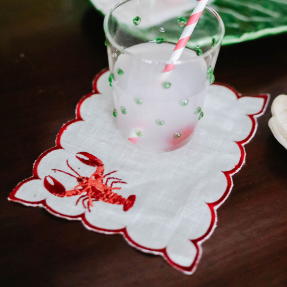 a white lobster napkin with an embroidered orange lobster motif and red scalloped edges with a glass resting on top