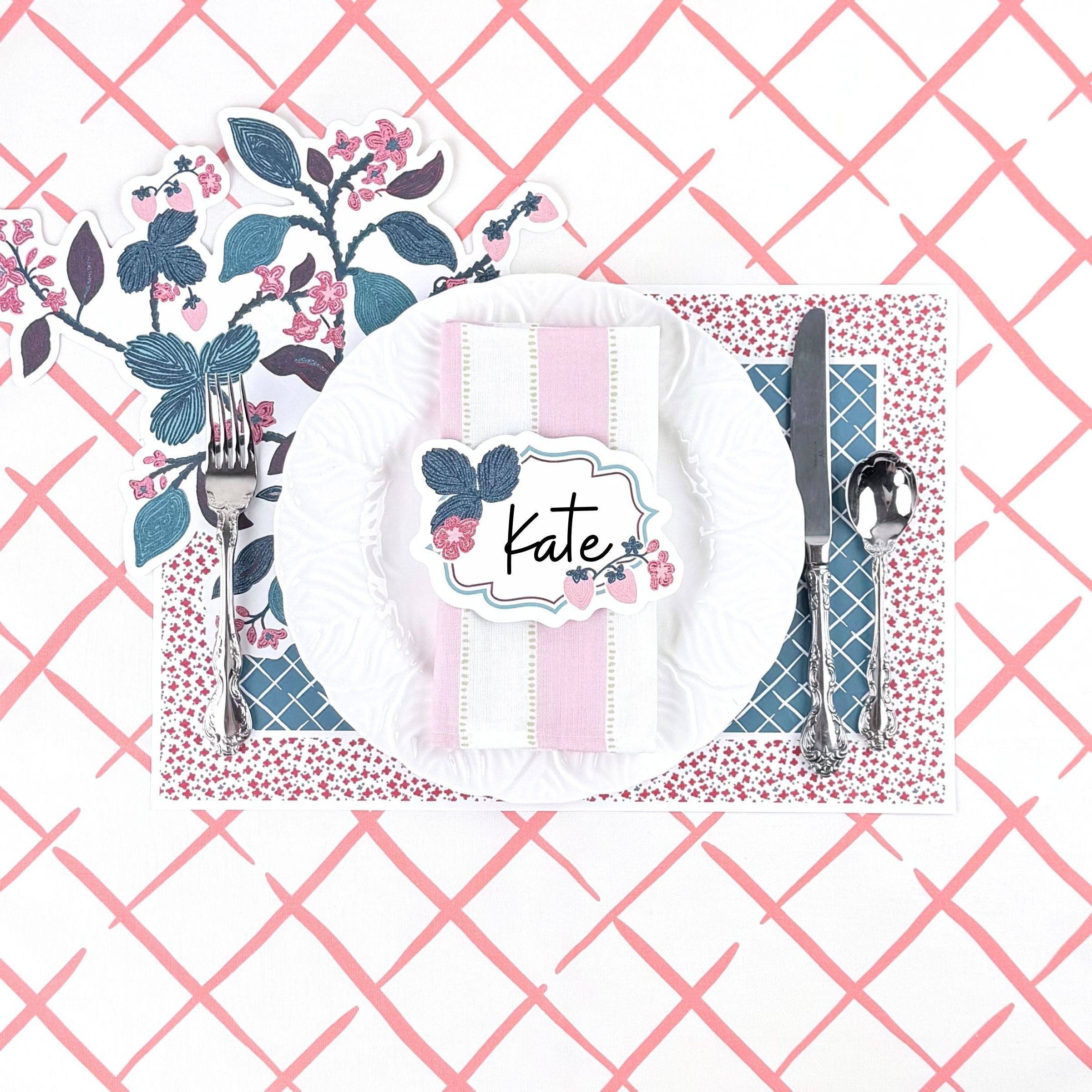 Floral strawberry themed table setting featuring a decorative place card, white dinner plate, patterned placemat, and pink trellis background.