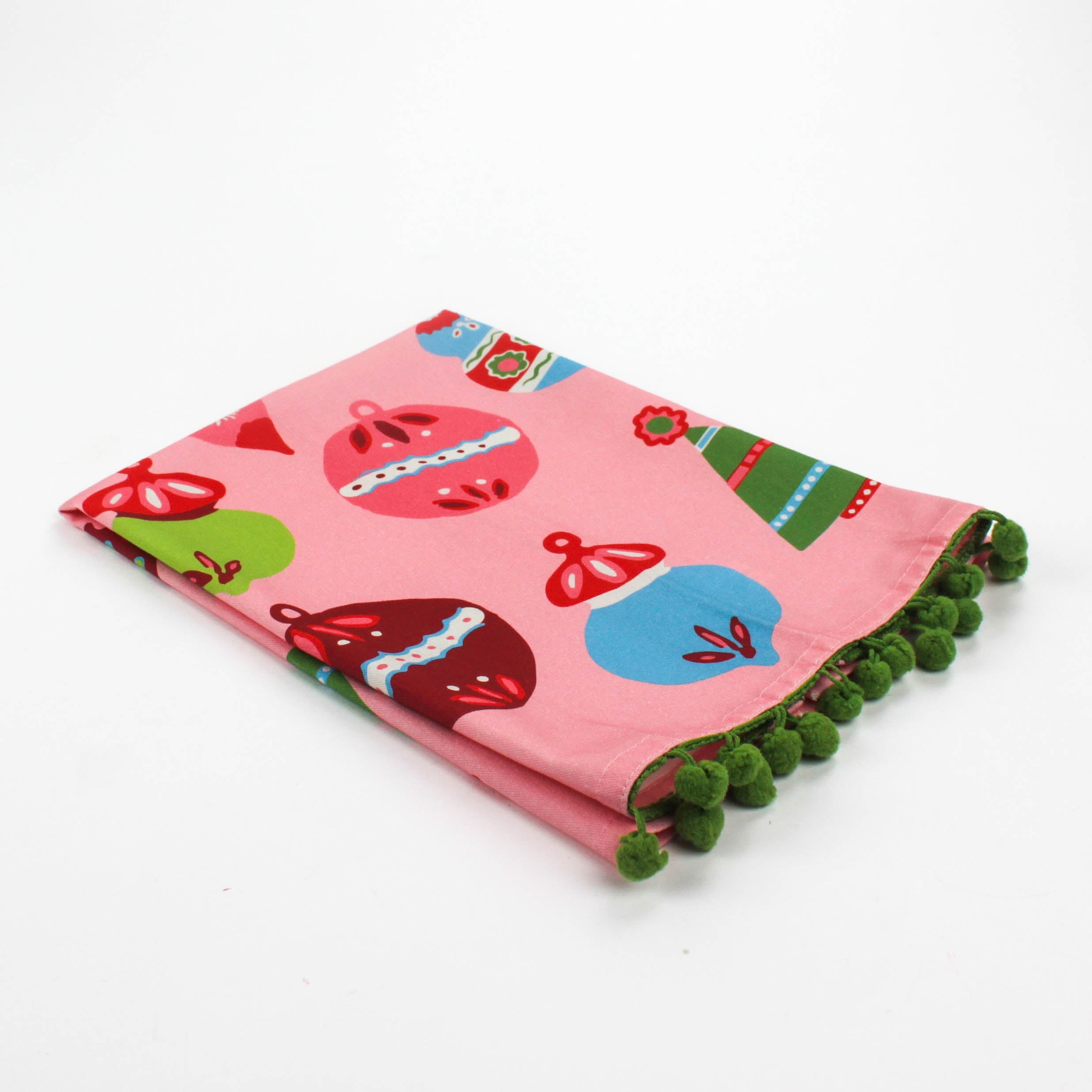 A folded pink christmas tea towel trimmed with green pom pom and colorful Christmas-themed decorations on a white background