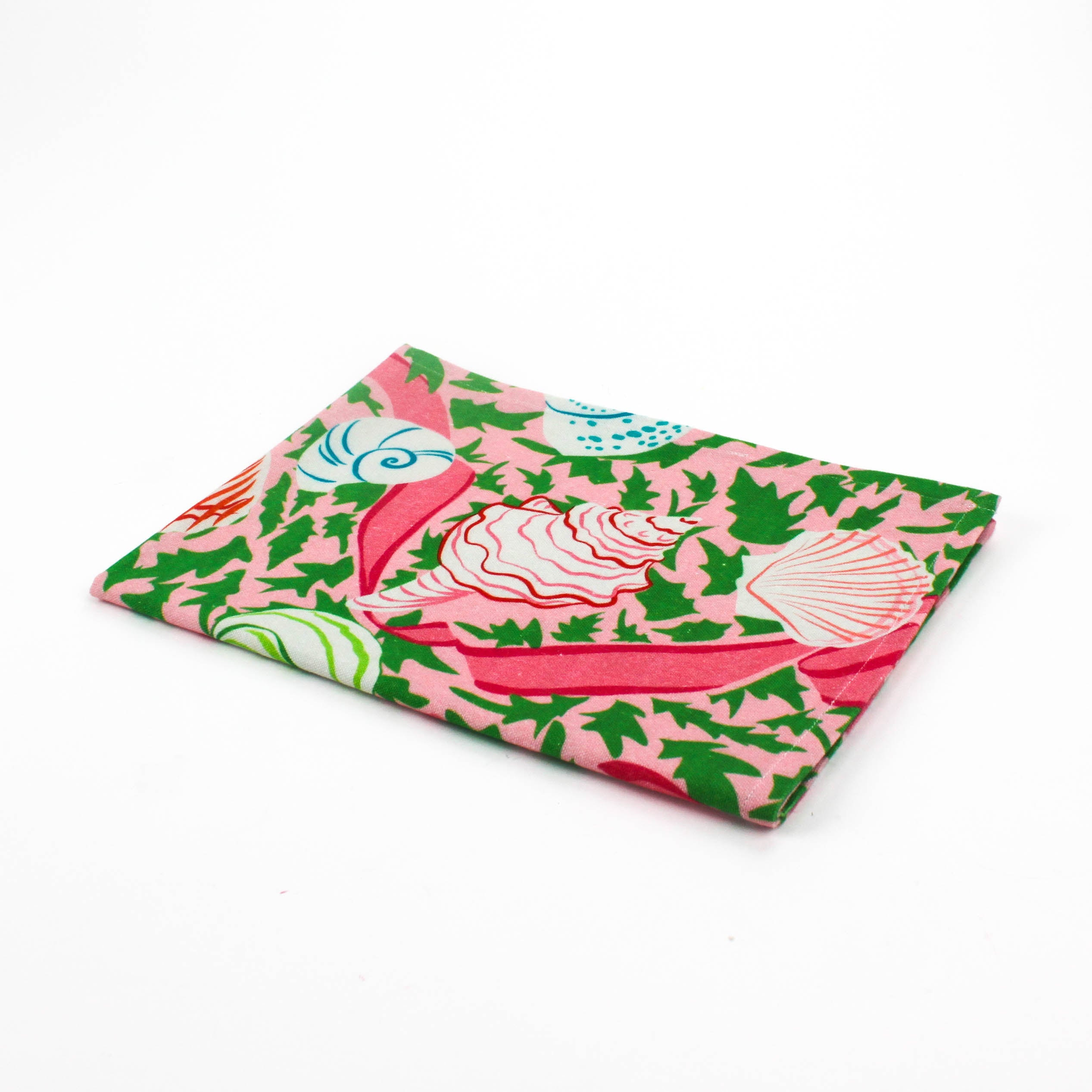 Folded Christmas tea towel featuring a pink background with colorful seashell and ribbon patterns accented by green holly-like leaves for a festive coastal look.