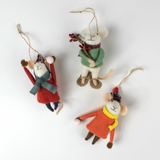 Three felt mouse ornaments dressed for winter—one in a red coat and plaid hat tossing a snowball, one in an orange coat with a yellow scarf holding a snowball, and one in a green sweater holding red berries—lying in white surface.
