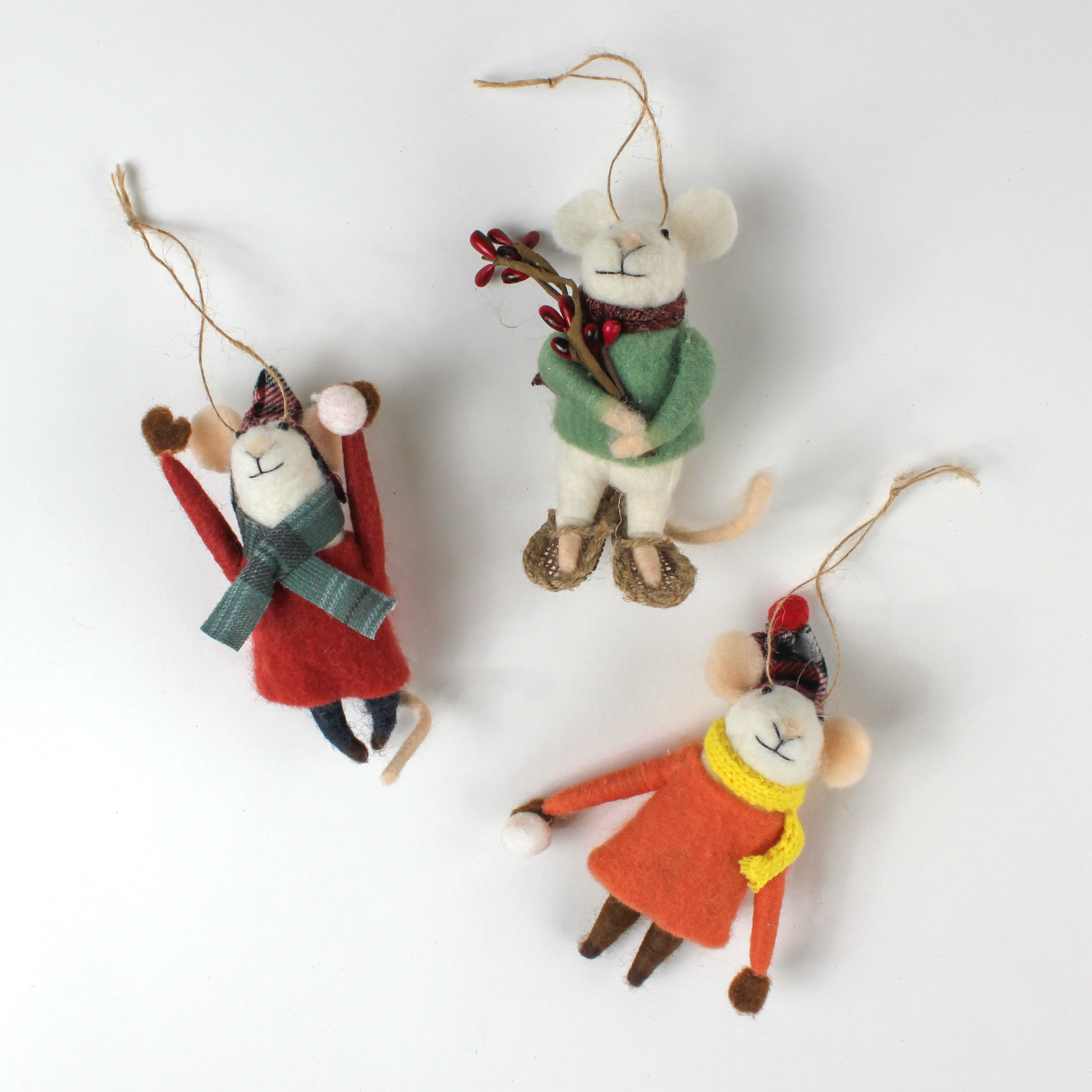 Three felt mouse ornaments dressed for winter—one in a red coat and plaid hat tossing a snowball, one in an orange coat with a yellow scarf holding a snowball, and one in a green sweater holding red berries—lying in white surface.
