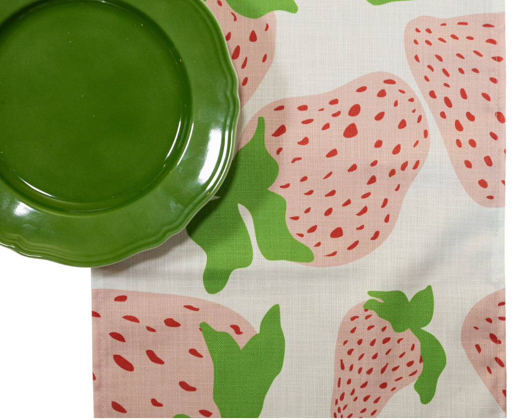 a Garden Party Strawberry cloth table runner designed by madcap cottage with motifs of large pink and green strawberries with green stem and a green plate resting on top