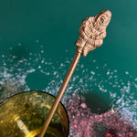 a close up of gilt metal gold christmas swizzle stick with decorative santa ends on a green background