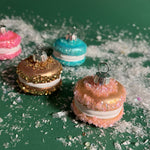 four glass macaron ornaments in brown, orange, pink and blue against a green snowy background. 