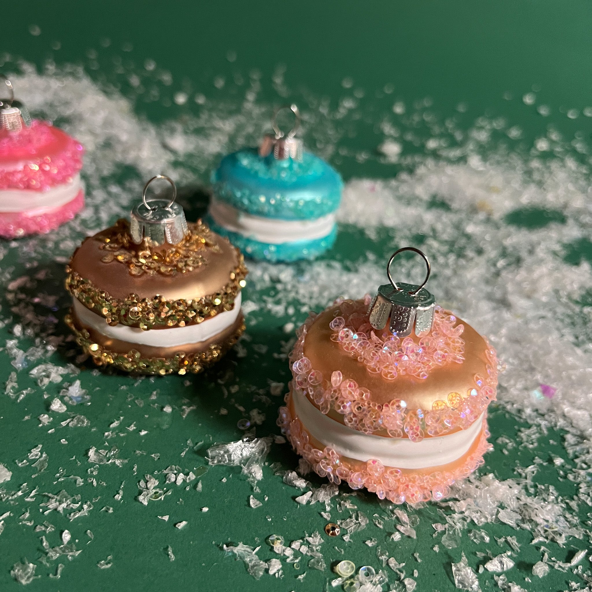 four glass macaron ornaments in brown, orange, pink and blue against a green snowy background. 