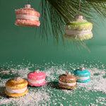 A set of six multicolored glass macaron ornaments hanging from a tree, each with a different color—pink, green, blue, beige, and yellow—with a glittery finish, placed on a bed of fake snow.