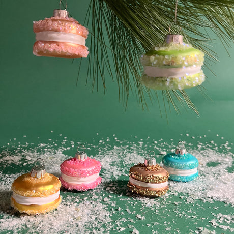 A set of six multicolored glass macaron ornaments hanging from a tree, each with a different color—pink, green, blue, beige, and yellow—with a glittery finish, placed on a bed of fake snow.