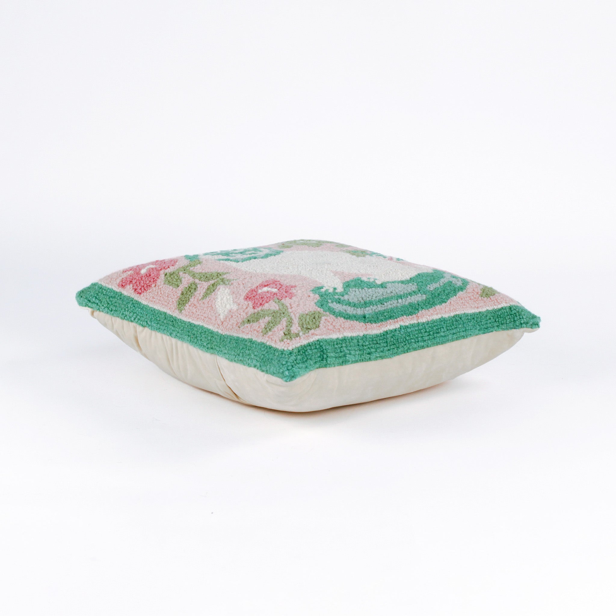 side view of a square decorative pillow with a hooked texture, featuring a white pug with green details surrounded by pink, white, and green floral accents on a blush pink background.