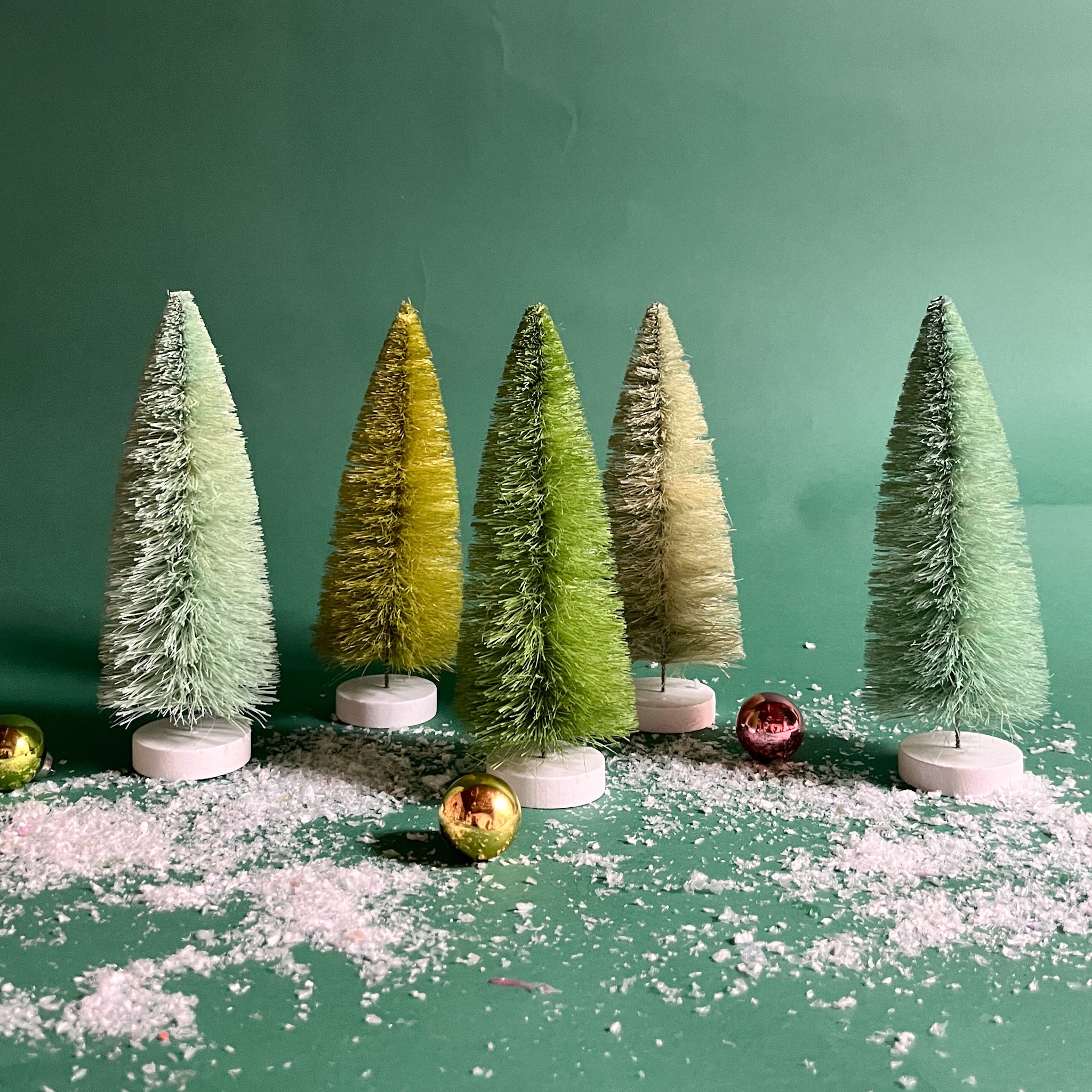 a set of 5, Green Bottle Brush Christmas Tree table decorations in different tones against a green, snow covered background. 