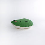 Frog-shaped decorative pillow with a hooked texture, featuring a smiling green frog face with pink cheeks and large black eyes lying in a white surface