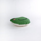 Frog-shaped decorative pillow with a hooked texture, featuring a smiling green frog face with pink cheeks and large black eyes lying in a white surface