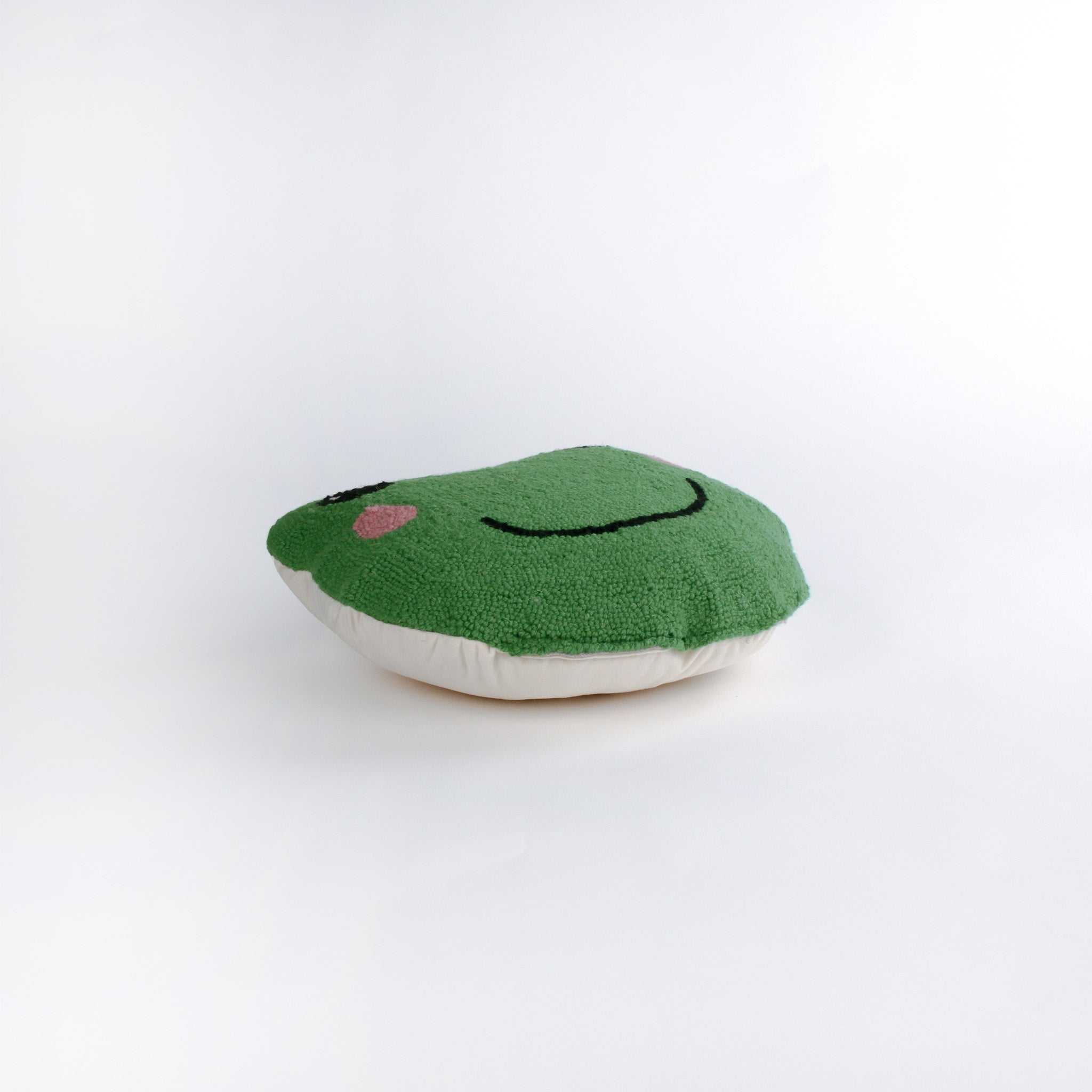 Frog-shaped decorative pillow with a hooked texture, featuring a smiling green frog face with pink cheeks and large black eyes lying in a white surface