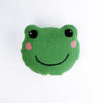 Frog-shaped decorative pillow with a hooked texture, featuring a smiling green frog face with pink cheeks and large black eyes.