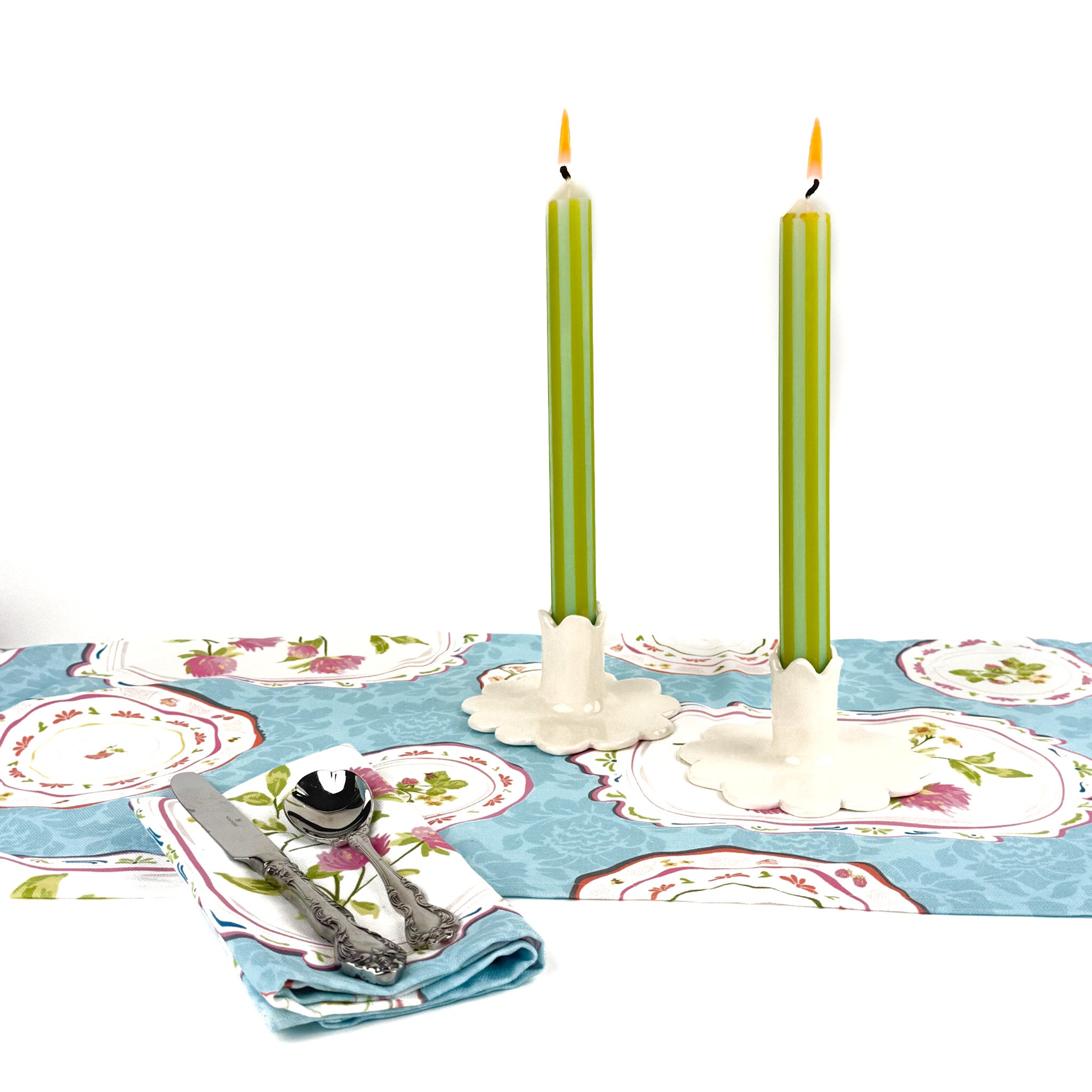 a pair of white scalloped candle holders with lighted green and white stripe candles on top of a spring themed table runner and a dinner napkin cloth