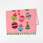 A pink christmas tea towel folded in half trimmed with green pom pom and colorful Christmas-themed decorations on a white background