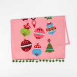 A pink christmas tea towel folded in half trimmed with green pom pom and colorful Christmas-themed decorations on a white background