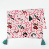 Pink Christmas tea towel featuring Nutcracker-themed illustrations with ribbons, ballerinas, swans, Christmas trees, and presents, finished with teal tassels on each corner.