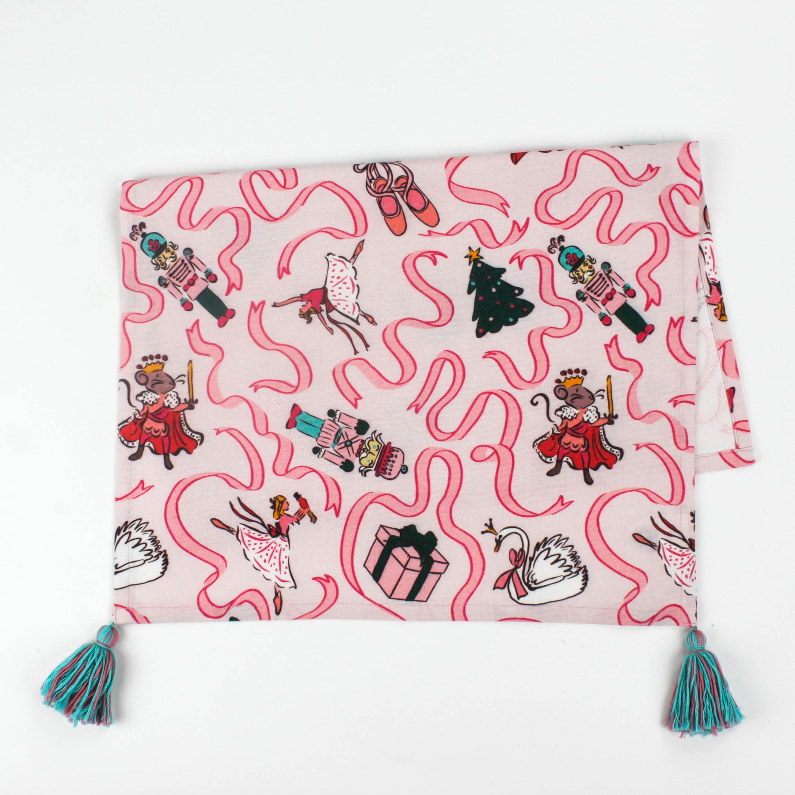 Pink Christmas tea towel featuring Nutcracker-themed illustrations with ribbons, ballerinas, swans, Christmas trees, and presents, finished with teal tassels on each corner.