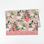 Folded Christmas tea towel with a pink background featuring a floral chinoiserie design and decorative pug dogs among green and red vases, bordered with a geometric pattern.