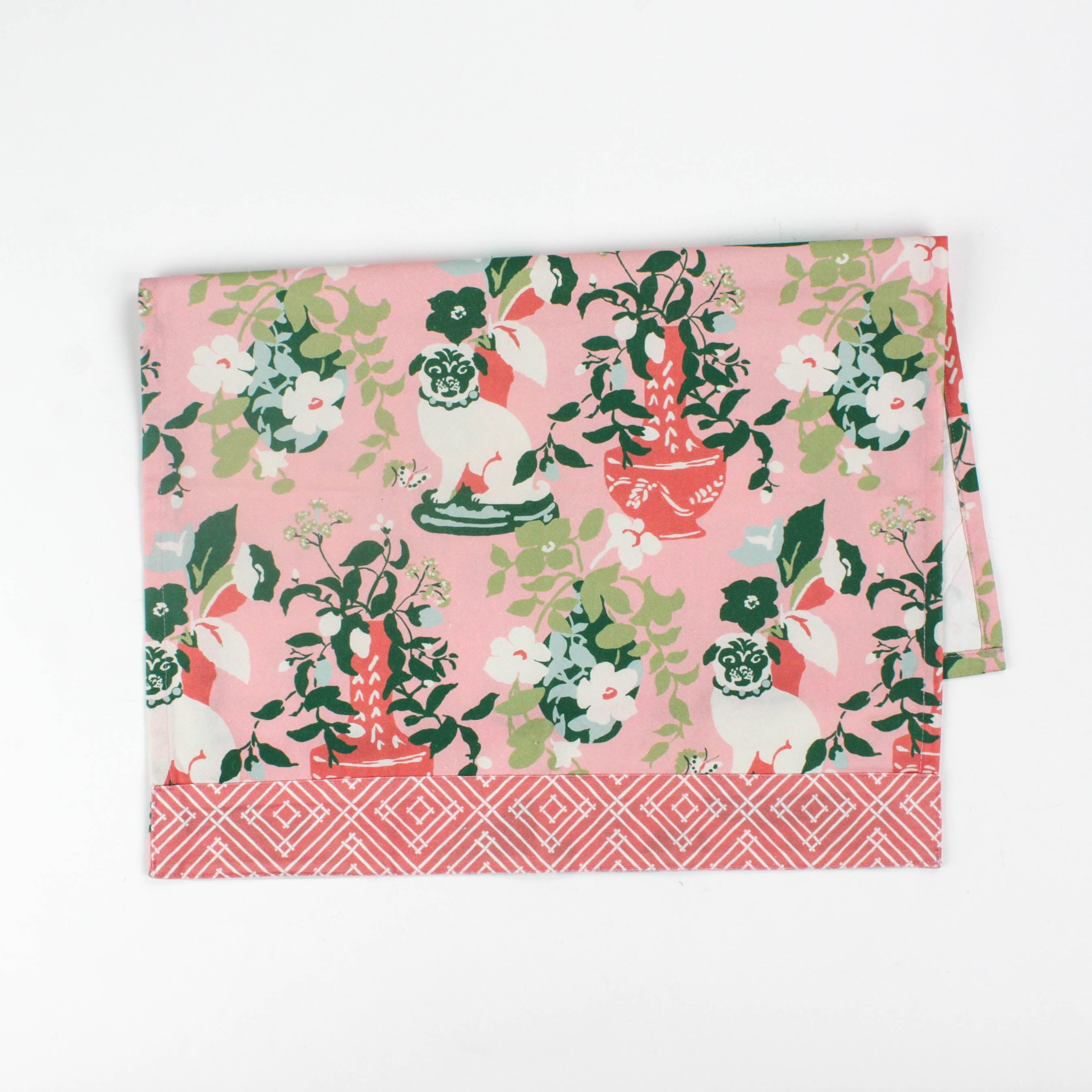 Folded Christmas tea towel with a pink background featuring a floral chinoiserie design and decorative pug dogs among green and red vases, bordered with a geometric pattern.