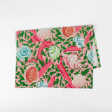 Folded Christmas tea towel featuring a pink background with colorful seashell and ribbon patterns accented by green holly-like leaves for a festive coastal look.