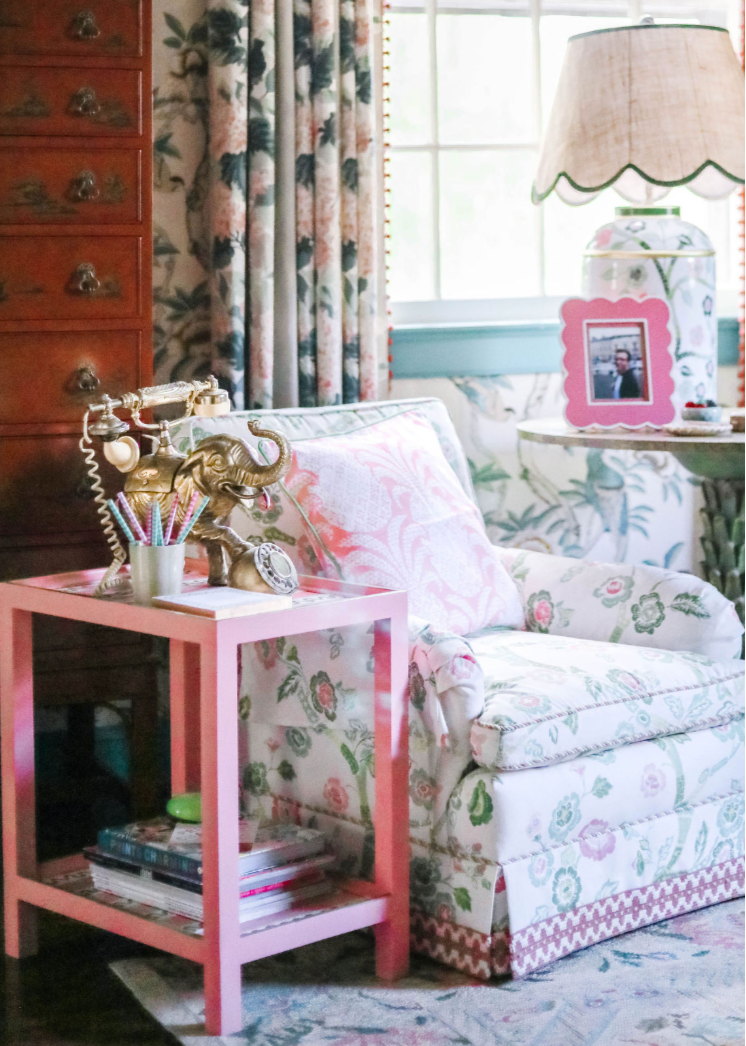 A pink side table with green and white striped accents and an elephant phone and notepad resting on top next to a chinoiserie fabric chair