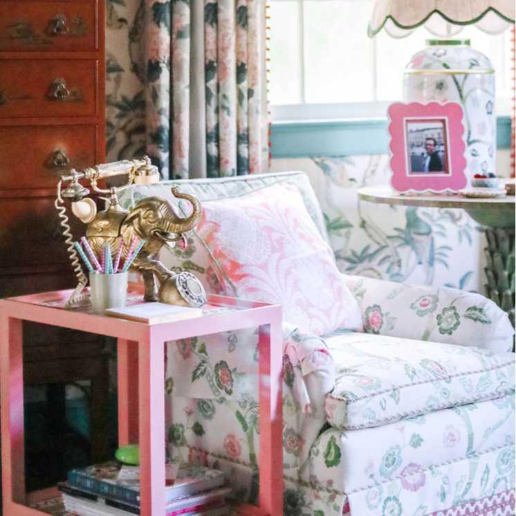 A pink side table with green and white striped accents and an elephant phone and notepad resting on top next to a chinoiserie fabric chair
