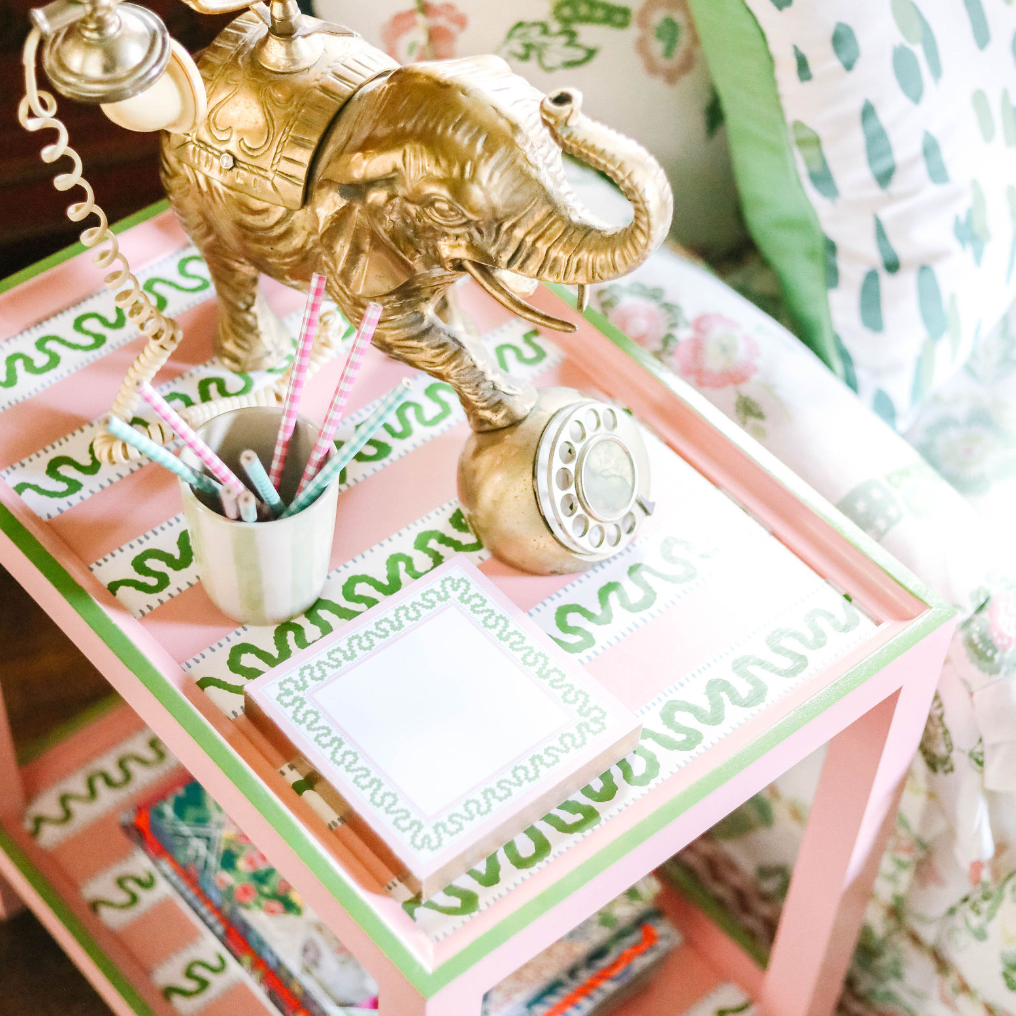 A pink side table with green and white striped accents and an elephant phone and notepad resting on top. 
