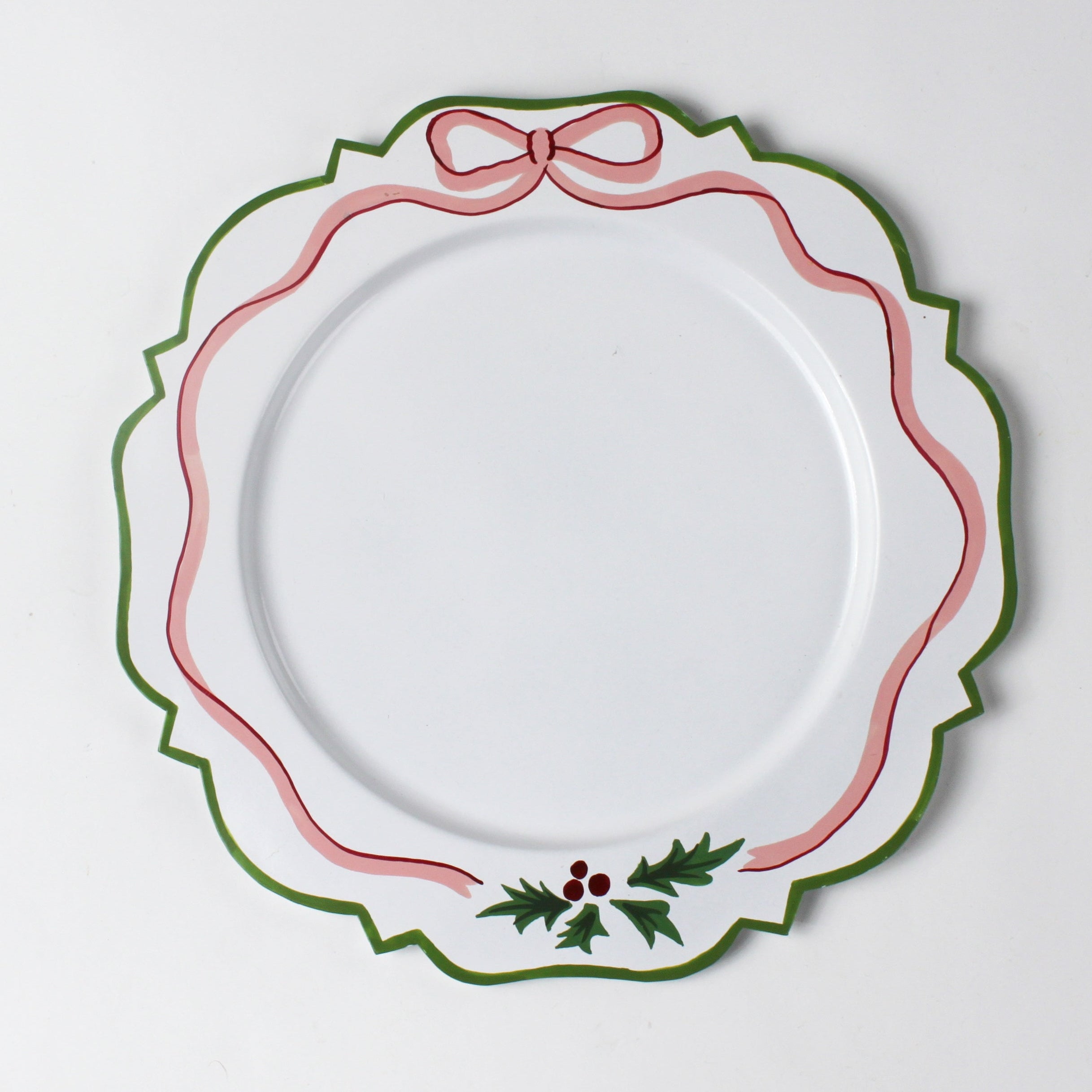 A white tole metal christmas charger with a pink ribbon and holly motifs around the edges against a white background.  