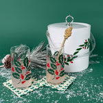 Two holly-printed holiday glasses with drinks beside a matching white holly ice bucket on a snowy green background