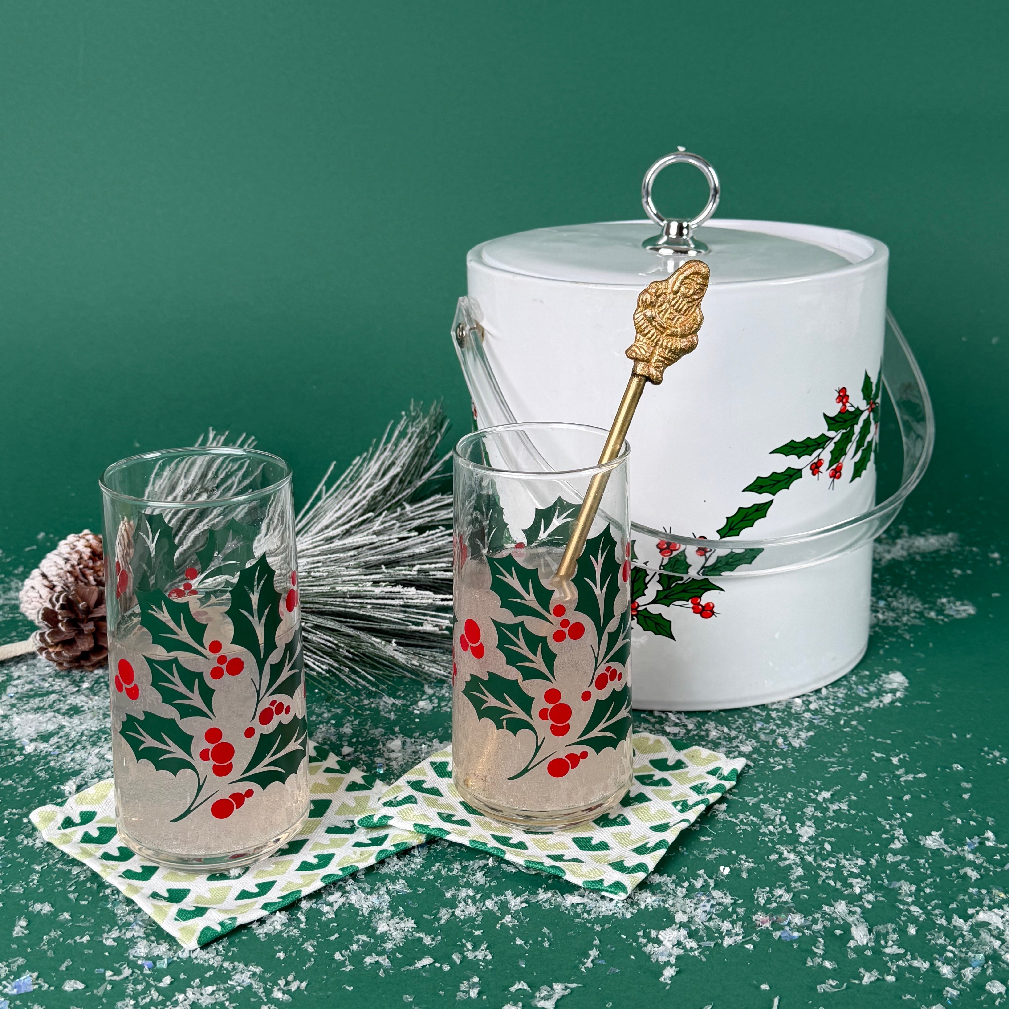 Two holly-printed holiday glasses with drinks beside a matching white holly ice bucket on a snowy green background