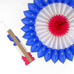 A folded red white and blue honeycomb paper fan decoration with an attached wooden support strip beside an opened patriotic fan on a white background.